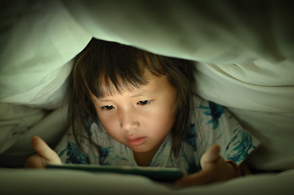 Child are watching video smart phone under the blanket on bed at night time light flashes reflected from the screen,children using games with addiction and cartoon concept .